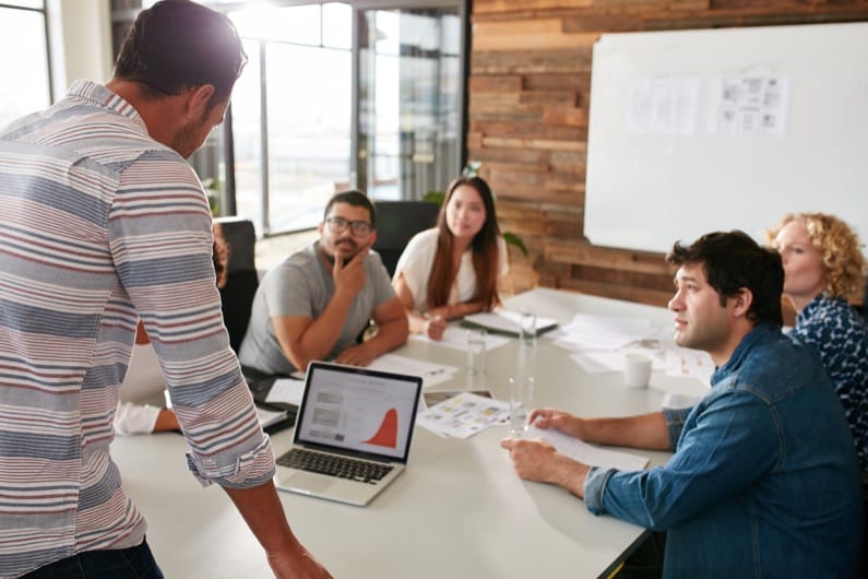 Young,Man,Giving,Business,Presentation,On,Laptop,To,Colleagues,Sitting Young man giving business presentation on laptop to colleagues sitting around table in conference room.